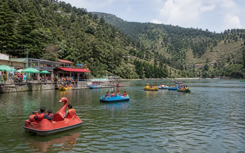 Families in life jackets enjoying paddle boats on a small hill lake near Mussoorie with forested slopes around.