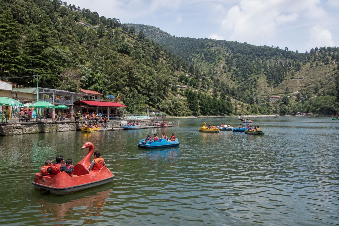 Families in life jackets enjoying paddle boats on a small hill lake near Mussoorie with forested slopes around.