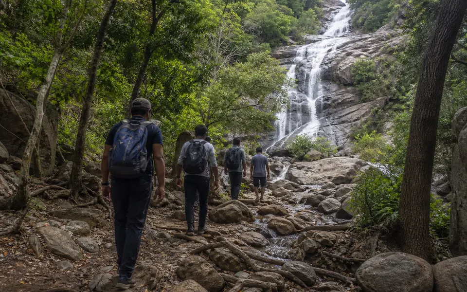 Trekker group walking on a rocky forest trail towards a multi-tiered waterfall in the Eastern Ghats near Srikalahasti.