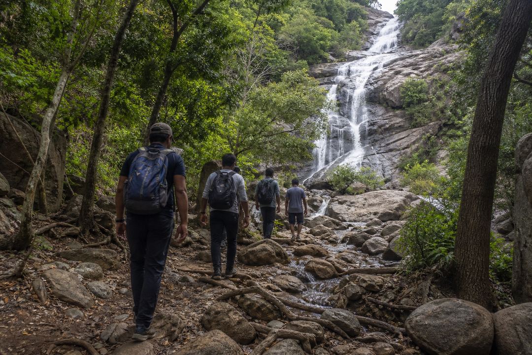 Trekker group walking on a rocky forest trail towards a multi-tiered waterfall in the Eastern Ghats near Srikalahasti.