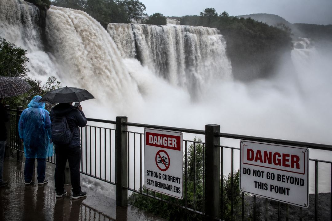 Indian waterfall in monsoon season with high water flow, safety barriers and warning signs, while visitors stand behind the railing.