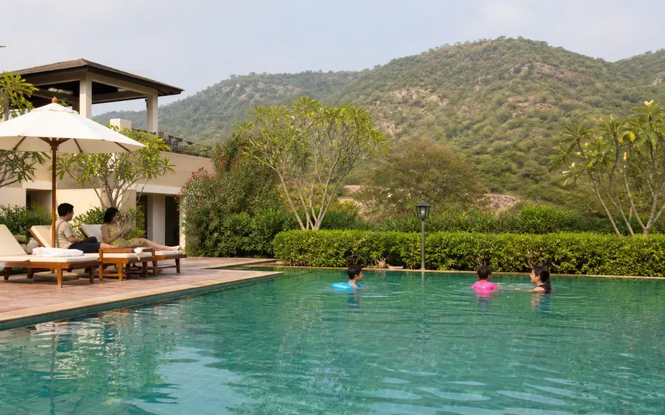Poolside area of a resort near Gurgaon with green Aravalli hills in the background and families relaxing by the water.