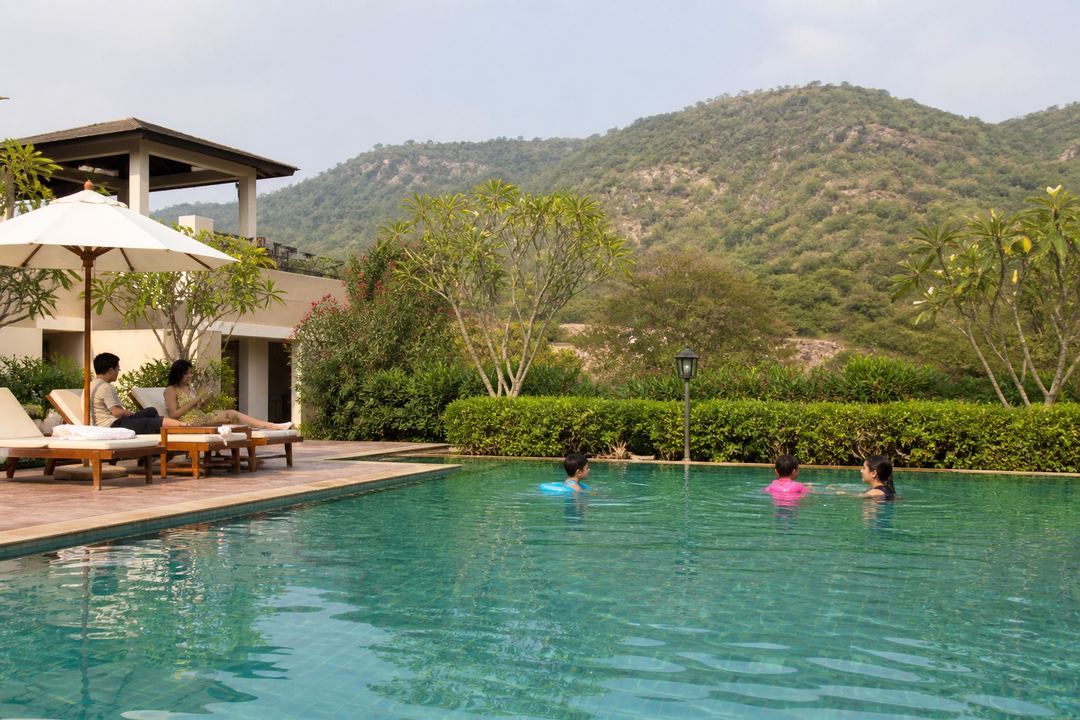 Poolside area of a resort near Gurgaon with green Aravalli hills in the background and families relaxing by the water.