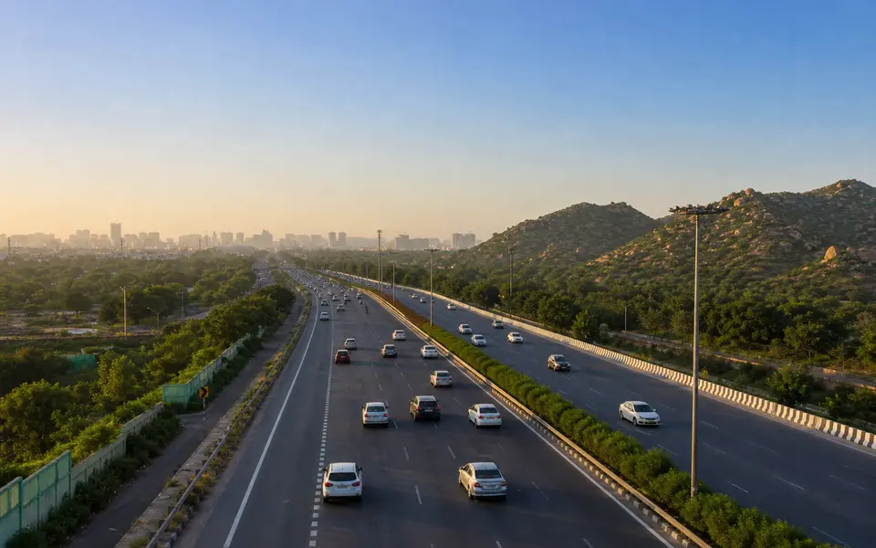 Cars leaving Gurgaon on a wide highway towards green Aravalli hills under a clear evening sky.