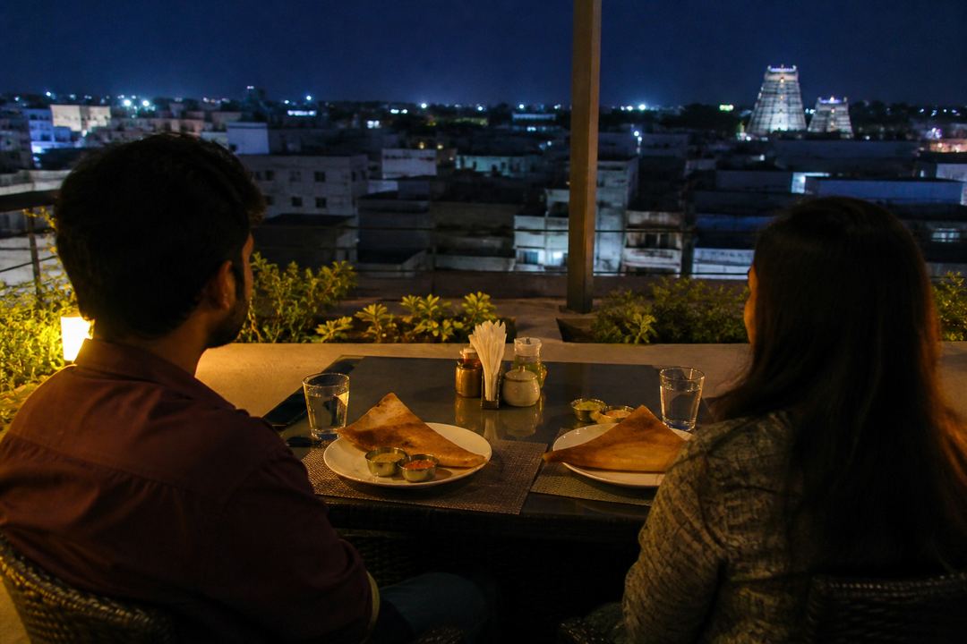 Rooftop restaurant table for two in Madurai at night, with simple South Indian dishes and city lights in the background.