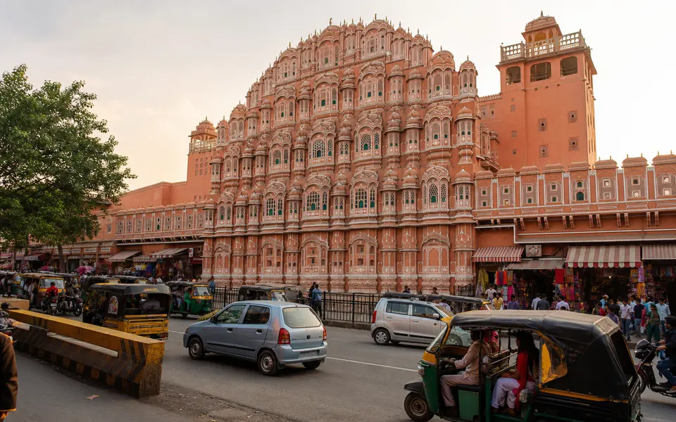 The pink sandstone façade of Hawa Mahal in Jaipur with auto-rickshaws, cars and shoppers moving along the busy street in front.