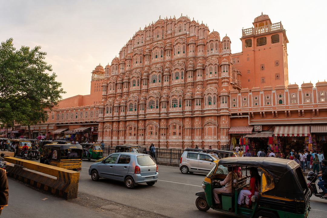 The pink sandstone façade of Hawa Mahal in Jaipur with auto-rickshaws, cars and shoppers moving along the busy street in front.