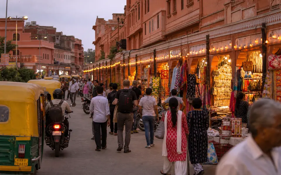 Evening scene in a Jaipur bazaar with painted pink buildings, arcaded shopfronts, and locals and visitors walking between stalls.