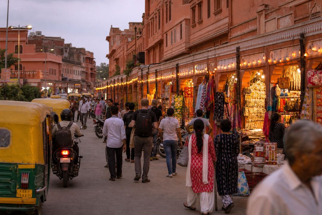 Evening scene in a Jaipur bazaar with painted pink buildings, arcaded shopfronts, and locals and visitors walking between stalls.