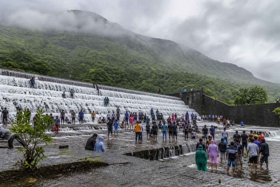 Monsoon water flowing over the steps of a dam near Lonavala with visitors standing at the edge and green hills behind.