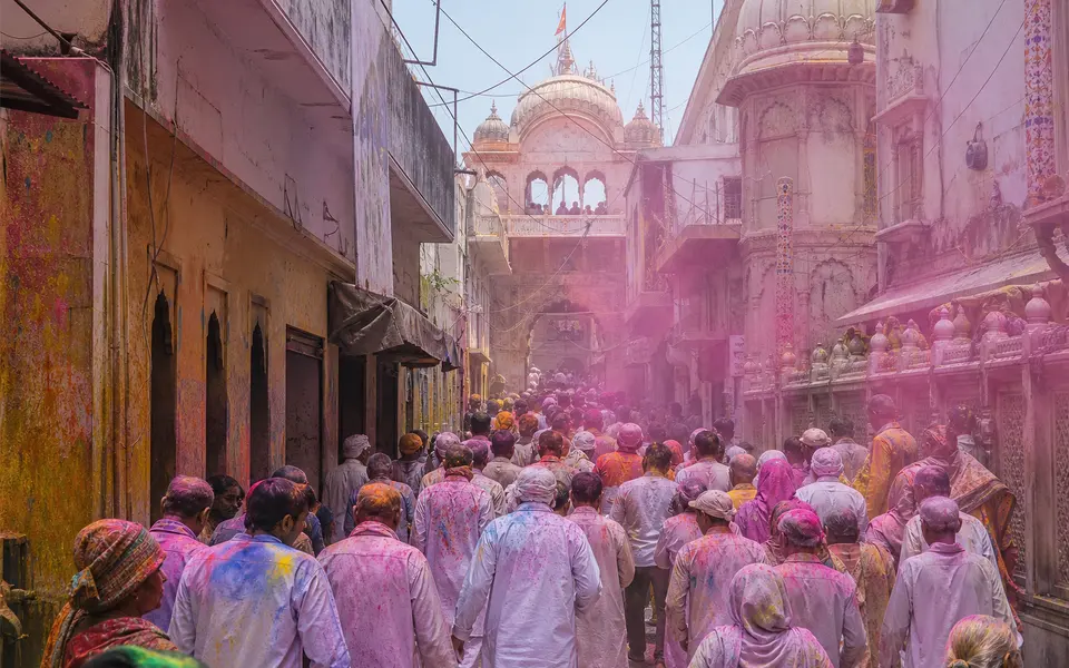 Street in Vrindavan during Holi with soft colours in the air, devotees in modest clothing, and a temple facade in the background.