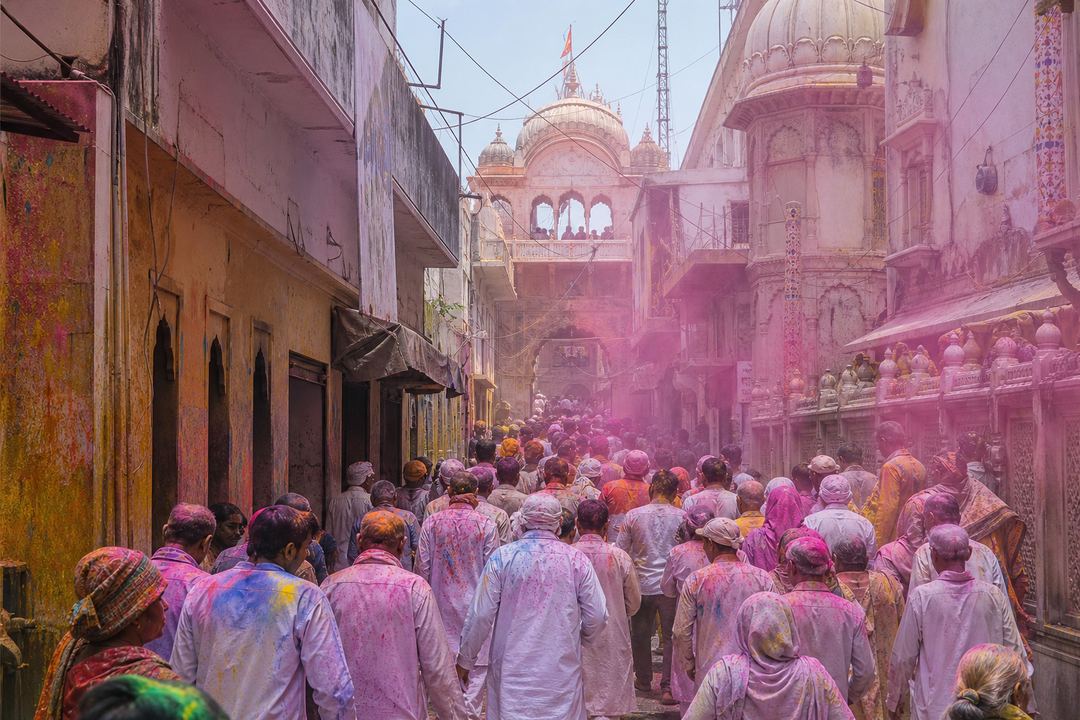 Street in Vrindavan during Holi with soft colours in the air, devotees in modest clothing, and a temple facade in the background.