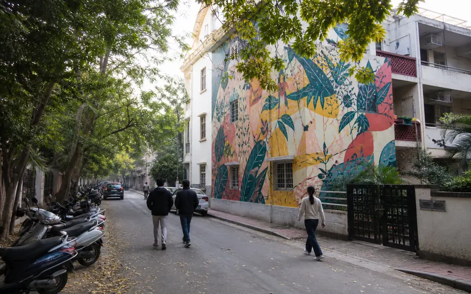 Colourful mural on a residential building in Lodhi Art District in Delhi, with a few walkers and parked scooters along a quiet street.