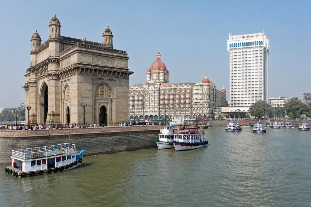 Gateway of India arch on Mumbai’s waterfront with boats in the harbour and heritage buildings behind it on a clear morning.