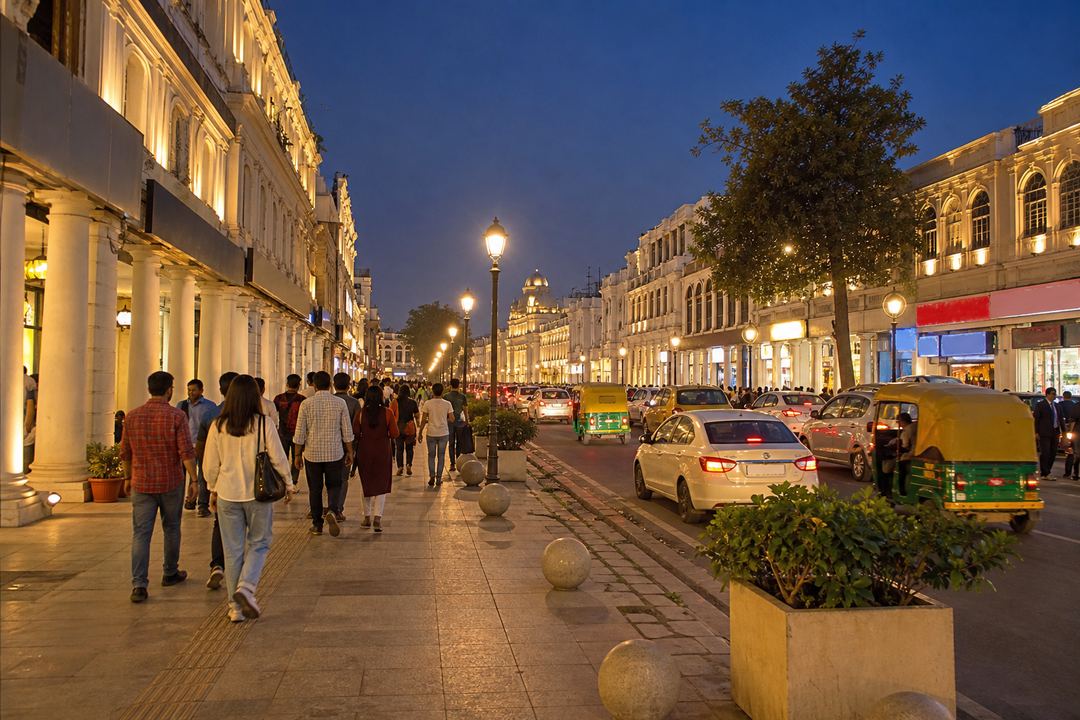 Evening view of Hazratganj in Lucknow with pedestrians, lit shopfronts and a line of cars along the road.