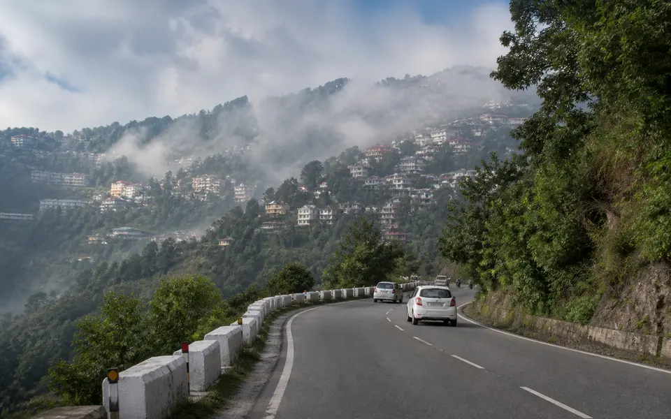 Curving mountain road leading to a small North Indian hill town with misty hills and houses on the slopes.