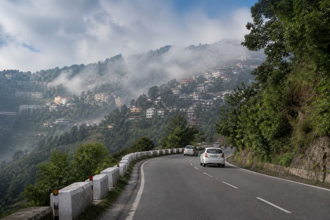 Curving mountain road leading to a small North Indian hill town with misty hills and houses on the slopes.