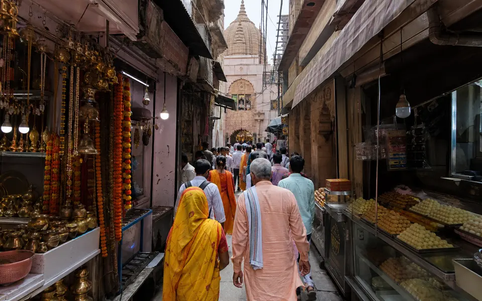 A narrow old-city lane in Varanasi with small shops, pilgrims and a view towards a temple entrance.