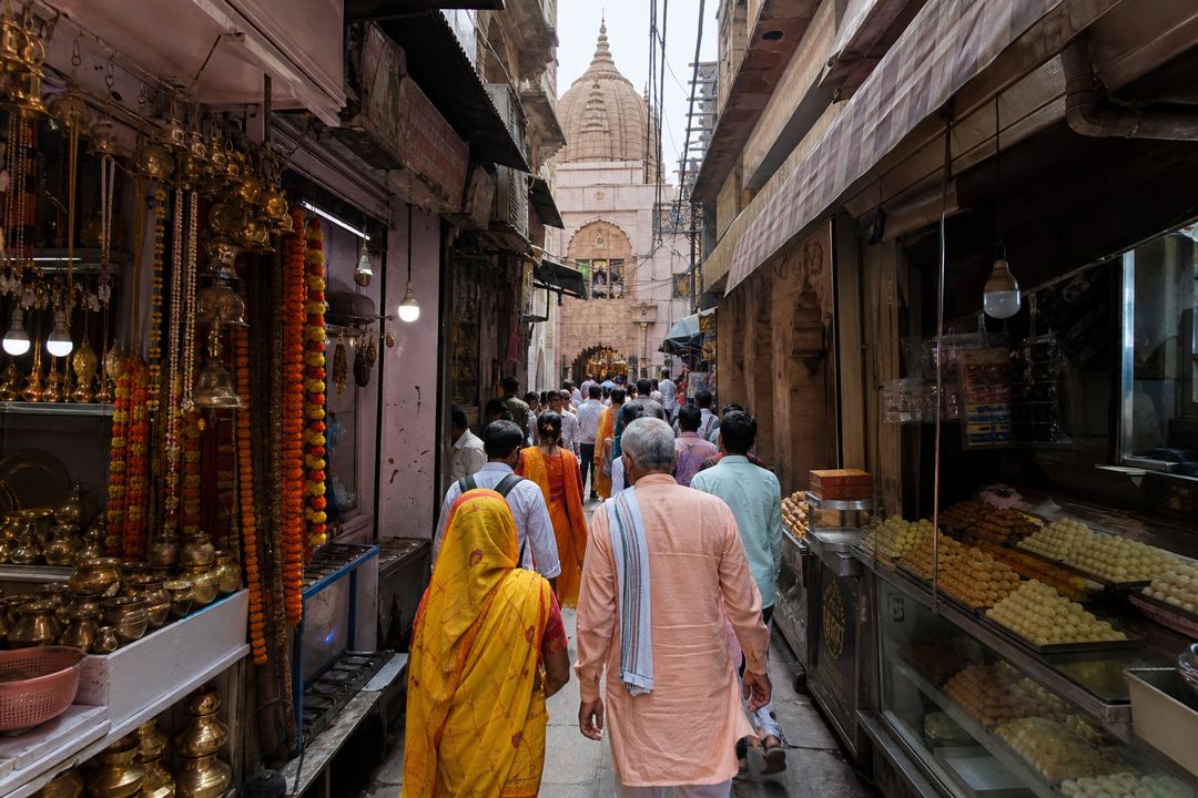 A narrow old-city lane in Varanasi with small shops, pilgrims and a view towards a temple entrance.