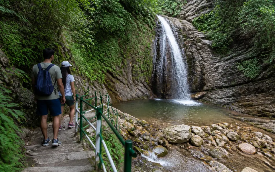 Short stone trail with steps leading through green forest to Neer Garh-style waterfall and small natural pool near Rishikesh, with a couple standing at a safe distance.