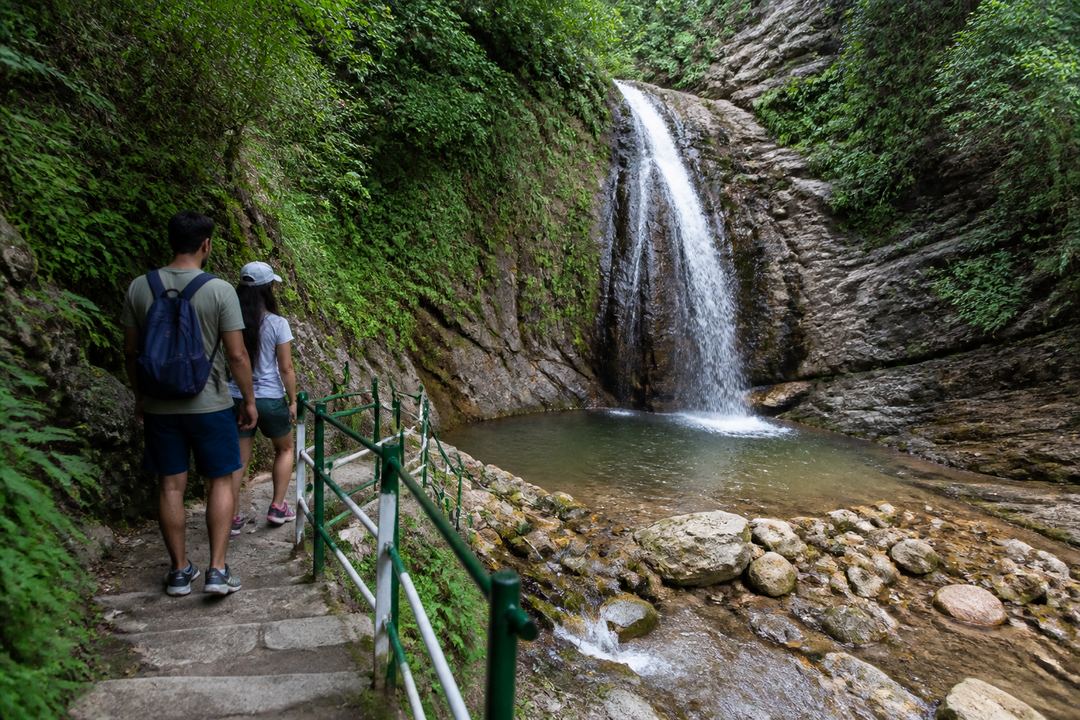Short stone trail with steps leading through green forest to Neer Garh-style waterfall and small natural pool near Rishikesh, with a couple standing at a safe distance.