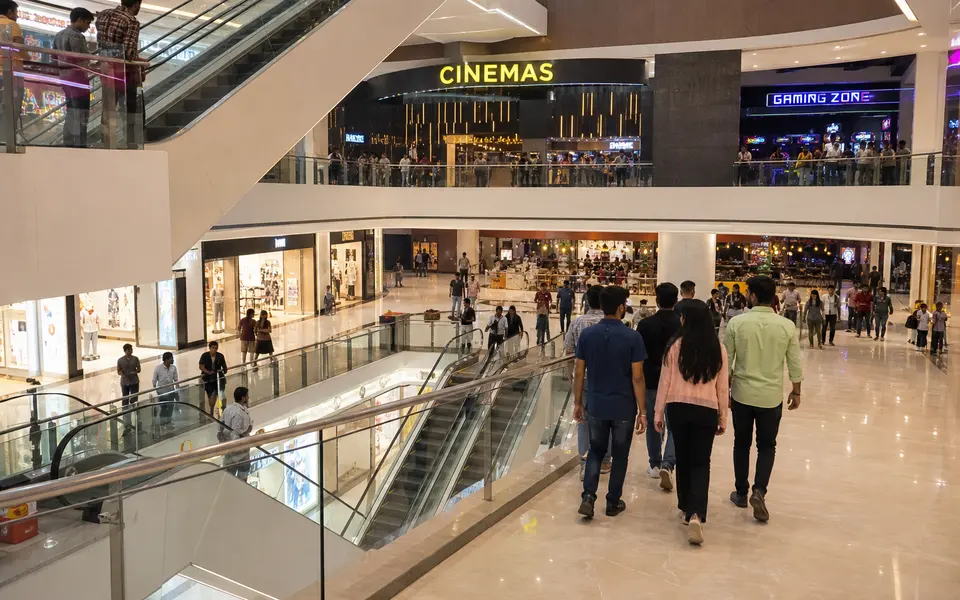 Interior of a large modern mall with escalators, a cinema sign and groups of young people walking between stores.