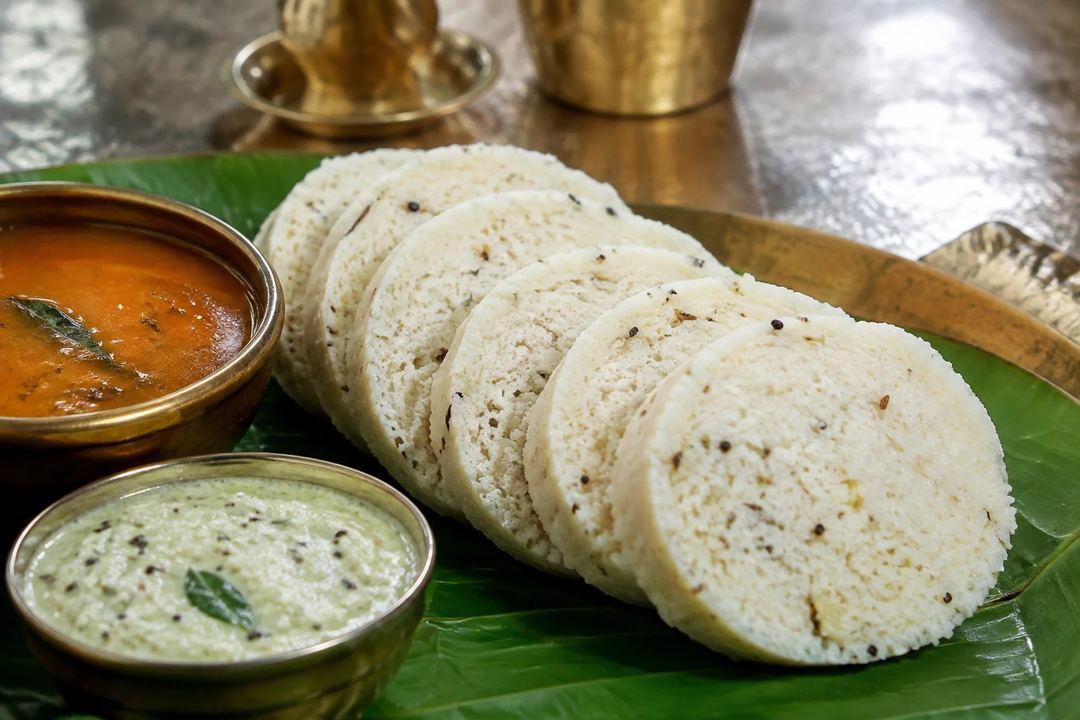 Thick round slices of seasoned Kanchipuram idli with visible pepper and cumin beside sambar and coconut chutney