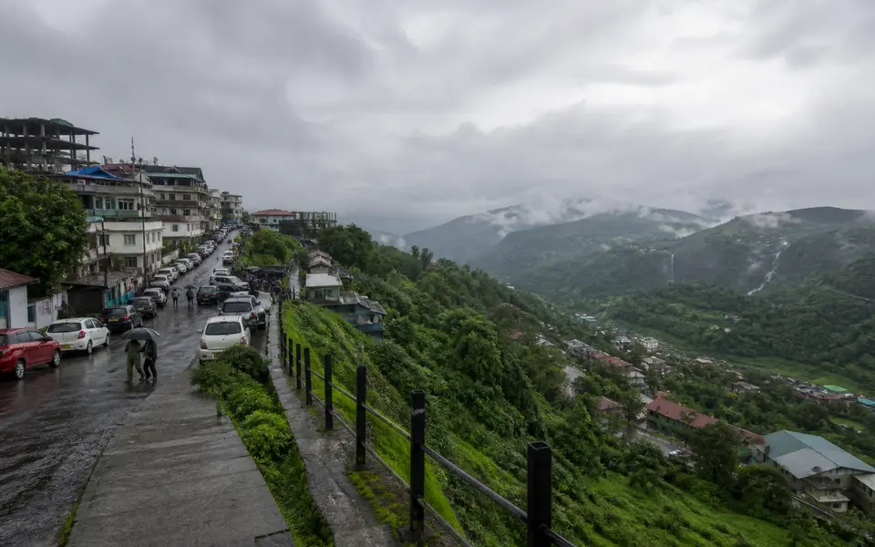 View of a lush Indian hill town and surrounding green valleys in light monsoon rain, seen from a safe, railed viewpoint with cloudy skies.