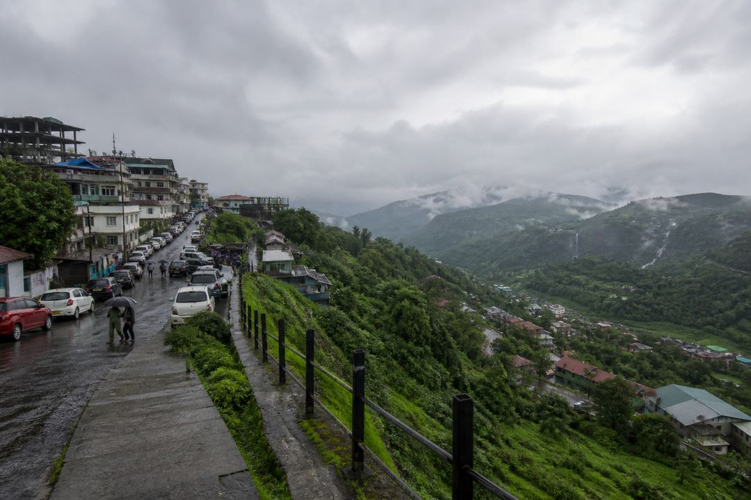 View of a lush Indian hill town and surrounding green valleys in light monsoon rain, seen from a safe, railed viewpoint with cloudy skies.
