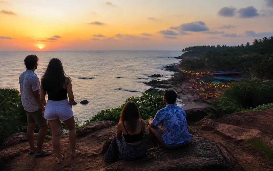 Group of friends at a rocky sunset viewpoint in North Goa, looking out over the sea with beach shacks lit below.