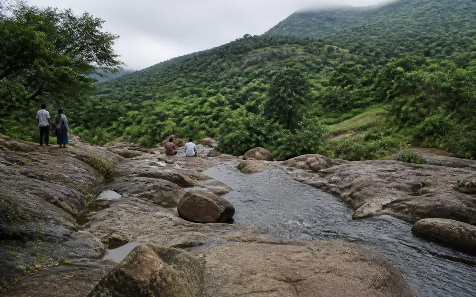 Green hills and a rocky stream near Puliancholai or Pachamalai with visitors standing at a safe distance from the water.