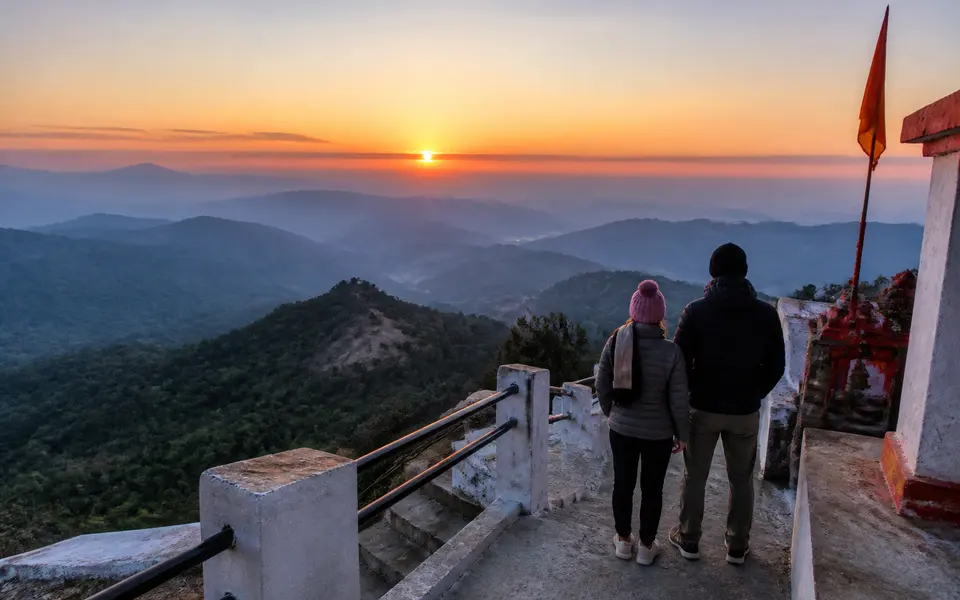 Sunrise view from a hilltop temple near Rishikesh with orange sky, layered foothills and a couple standing quietly at the viewpoint steps.