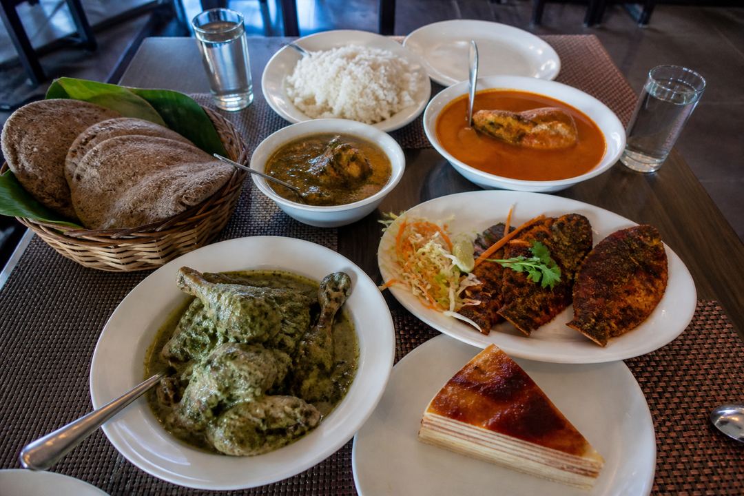 Casual Goan meal spread with fish curry, rice, fried fish, poi bread and bebinca dessert on a table in a simple restaurant.