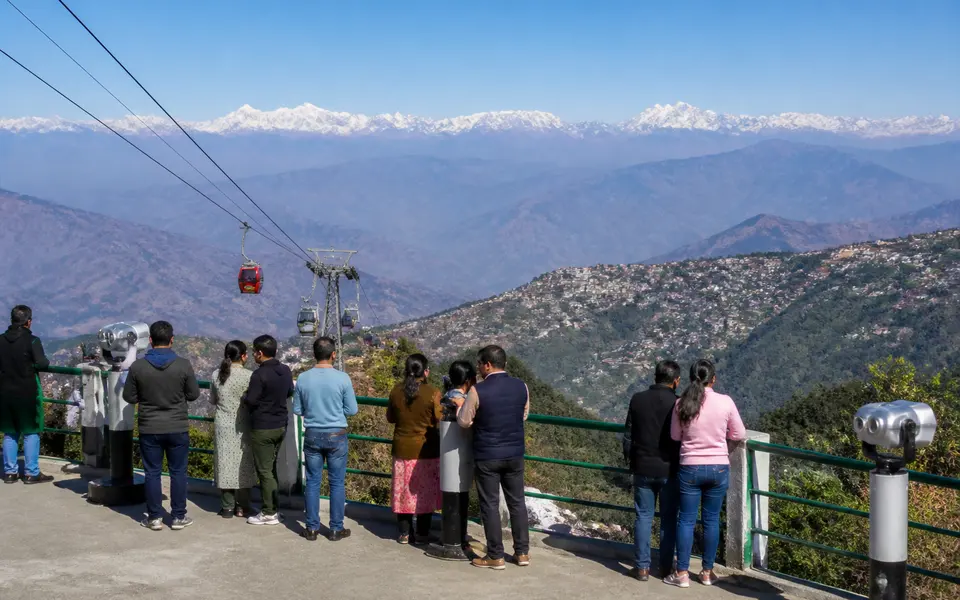 Tourists on a Mussoorie viewpoint terrace looking out over distant Himalayan peaks and the valley below.