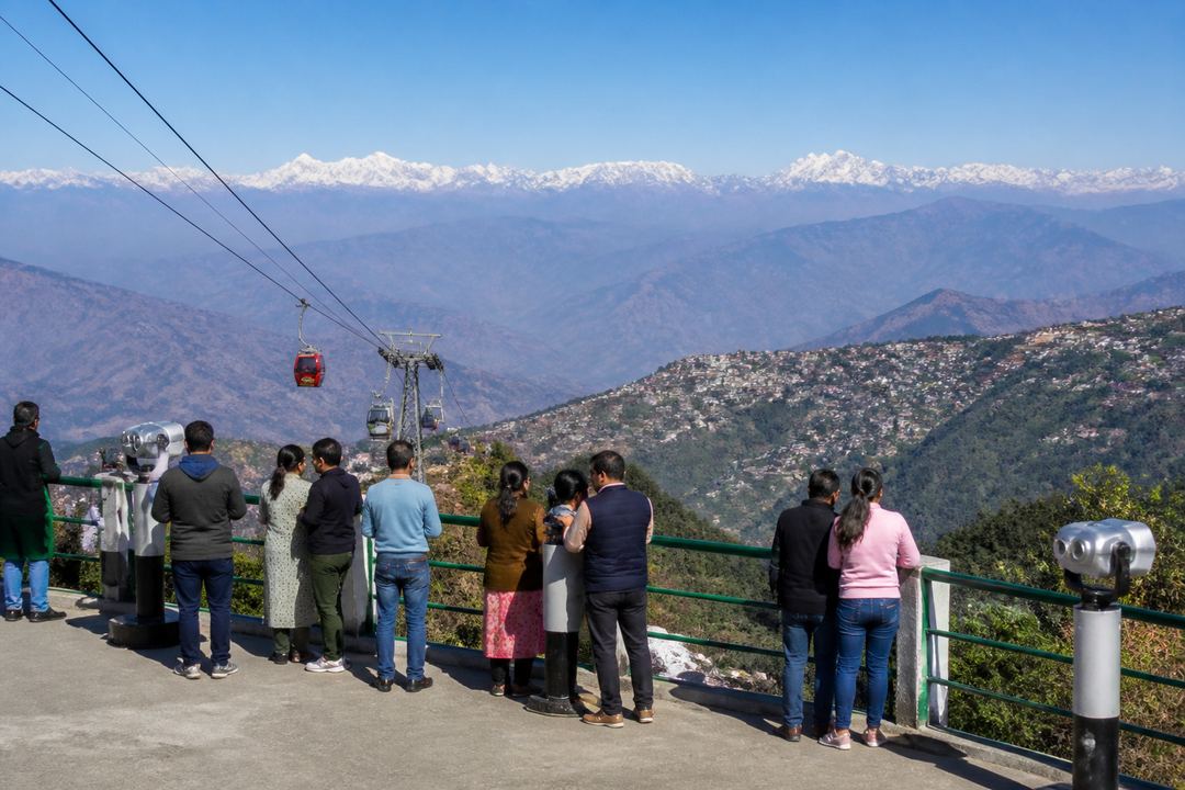 Tourists on a Mussoorie viewpoint terrace looking out over distant Himalayan peaks and the valley below.