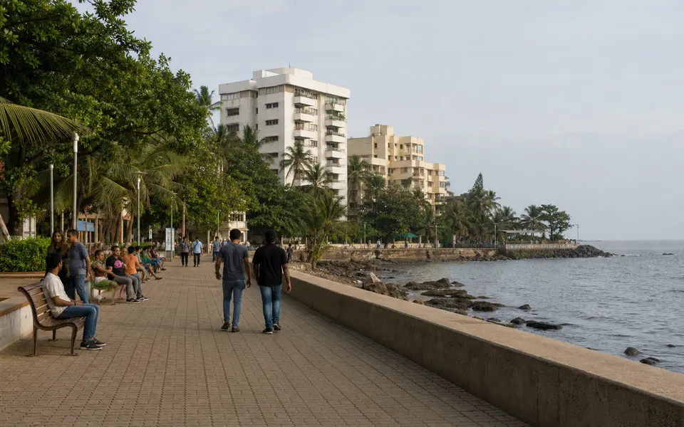 Bandra Bandstand promenade with people walking along the seafront, benches, and residential buildings in the background.
