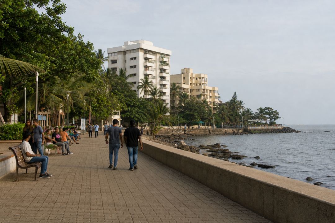 Bandra Bandstand promenade with people walking along the seafront, benches, and residential buildings in the background.