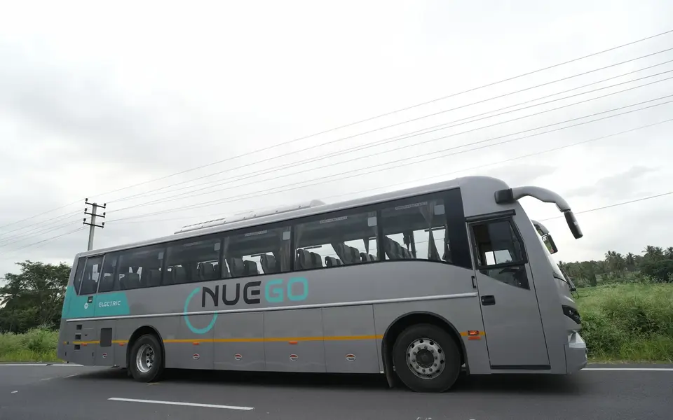 Silver and teal NueGo electric intercity coach photographed from the roadside while travelling on a divided highway.