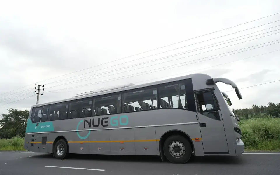 Side view of a silver and teal NueGo electric intercity coach on a road under a cloudy sky.