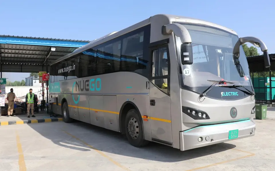 A silver and teal NueGo electric intercity coach parked at a boarding facility in daylight.