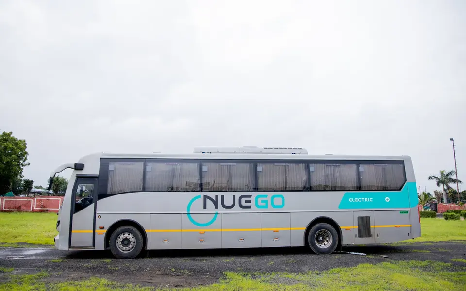 Full side profile of a silver and teal NueGo electric intercity coach, showing the body, wheels, luggage bays and roof equipment.