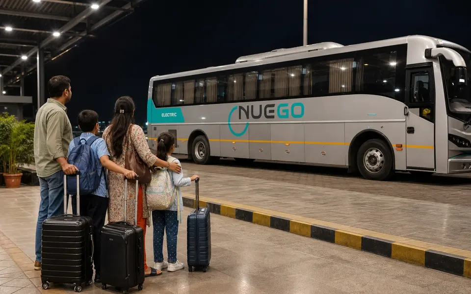 A family waits together with luggage in a well-lit terminal at night while a NueGo coach stands at the bay.