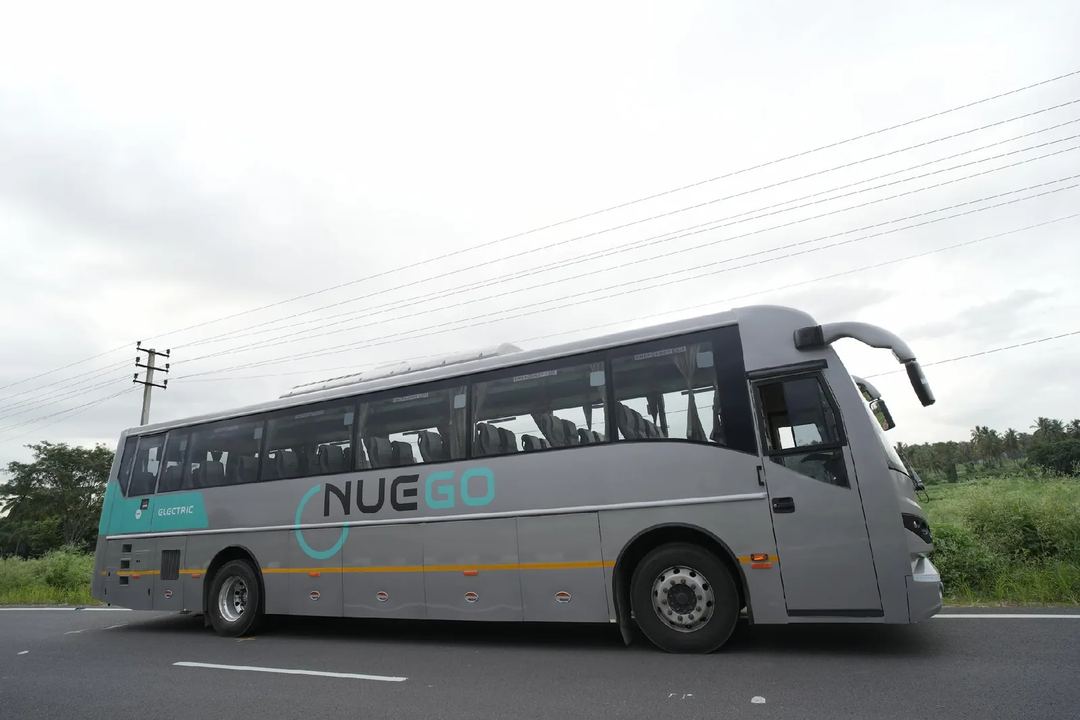 Side view of a silver and teal NueGo electric intercity coach on a road under a cloudy sky.