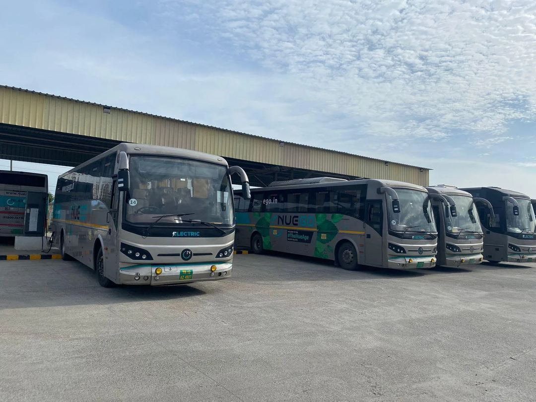 Several silver and teal NueGo electric intercity coaches parked in a depot under a blue sky.