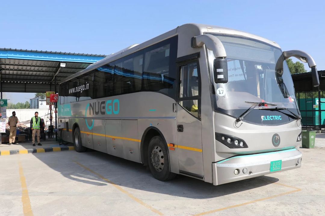A silver and teal NueGo electric intercity coach parked at a boarding facility in daylight.