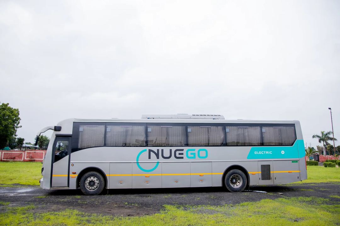 Full side view of a silver NueGo electric intercity coach with teal branding on a wet road surface.